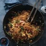 Stir fried thin noodles, chunks of meat, and green vegetables are in a black wok on a blue tablecloth. Green onion garnish sits beside the wok.