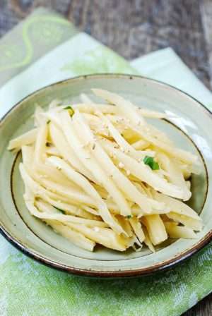 A pile of thinly sliced creamy white root vegetables sit in a grey clay bowl, garnished with sesame seeds and green onions.