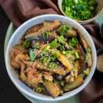 An ornate white bowl sits on a brown cloth napkin, filled with eggplant that has been cut lengthways and steamed, and garnished with green onions, chili flakes and sesame seeds.