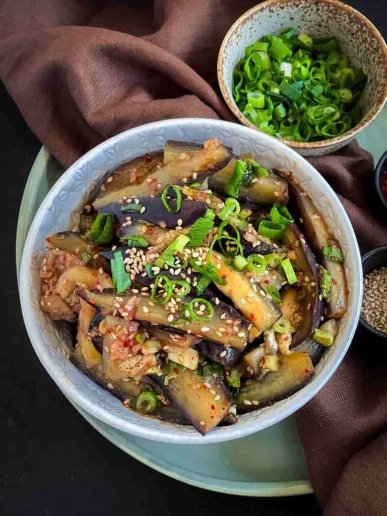 An ornate white bowl sits on a brown cloth napkin, filled with eggplant that has been cut lengthways and steamed, and garnished with green onions, chili flakes and sesame seeds.