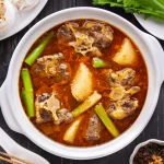 A white bowl sits on a brown table. The bowl is filled with a rich-looking red broth, stewed oxtail pieces, potatoes, and various green vegetables.