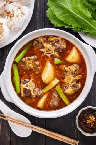 A white bowl sits on a brown table. The bowl is filled with a rich-looking red broth, stewed oxtail pieces, potatoes, and various green vegetables.