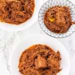 Shredded juicy red beef is piled in three white bowls on a white table.