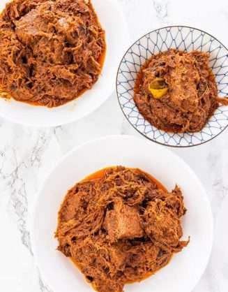 Shredded juicy red beef is piled in three white bowls on a white table.
