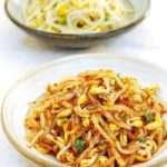 Two bowls filled with soybean sprouts on a white table. The one in the foreground is covered in a spicy dressing, and the one in the background is plain.