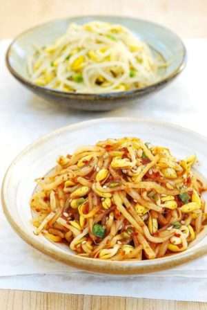 Two bowls filled with soybean sprouts on a white table. The one in the foreground is covered in a spicy dressing, and the one in the background is plain.