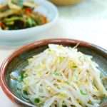 Blanched white mung bean sprouts are piled in a red and dark grey clay bowl on a white background.