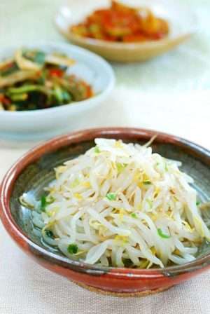 Blanched white mung bean sprouts are piled in a red and dark grey clay bowl on a white background.