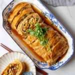 On a blue and white rectangular plate sits a meaty-looking golden sliced roll of tofu skin and vegetables, garnished with a sprig of herbs.