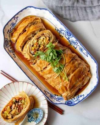 On a blue and white rectangular plate sits a meaty-looking golden sliced roll of tofu skin and vegetables, garnished with a sprig of herbs.