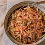 A clay bowl on a white tablecloth contains shredded chicken, chili sauce, sesame seeds and green cucumbers.