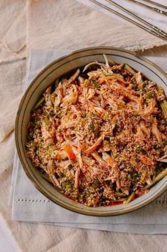 A clay bowl on a white tablecloth contains shredded chicken, chili sauce, sesame seeds and green cucumbers.