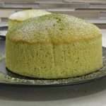 An unfrosted green soufflé cheesecake sits on a dark grey plate, lightly sprinkled with powdered sugar.