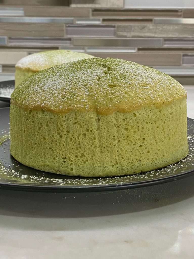 An unfrosted green soufflé cheesecake sits on a dark grey plate, lightly sprinkled with powdered sugar.